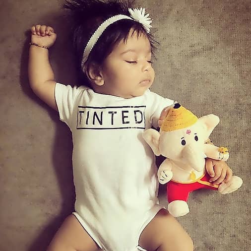 Baby wearing a 'TINTED' onesie holding a Ganesha plush toy against a neutral background