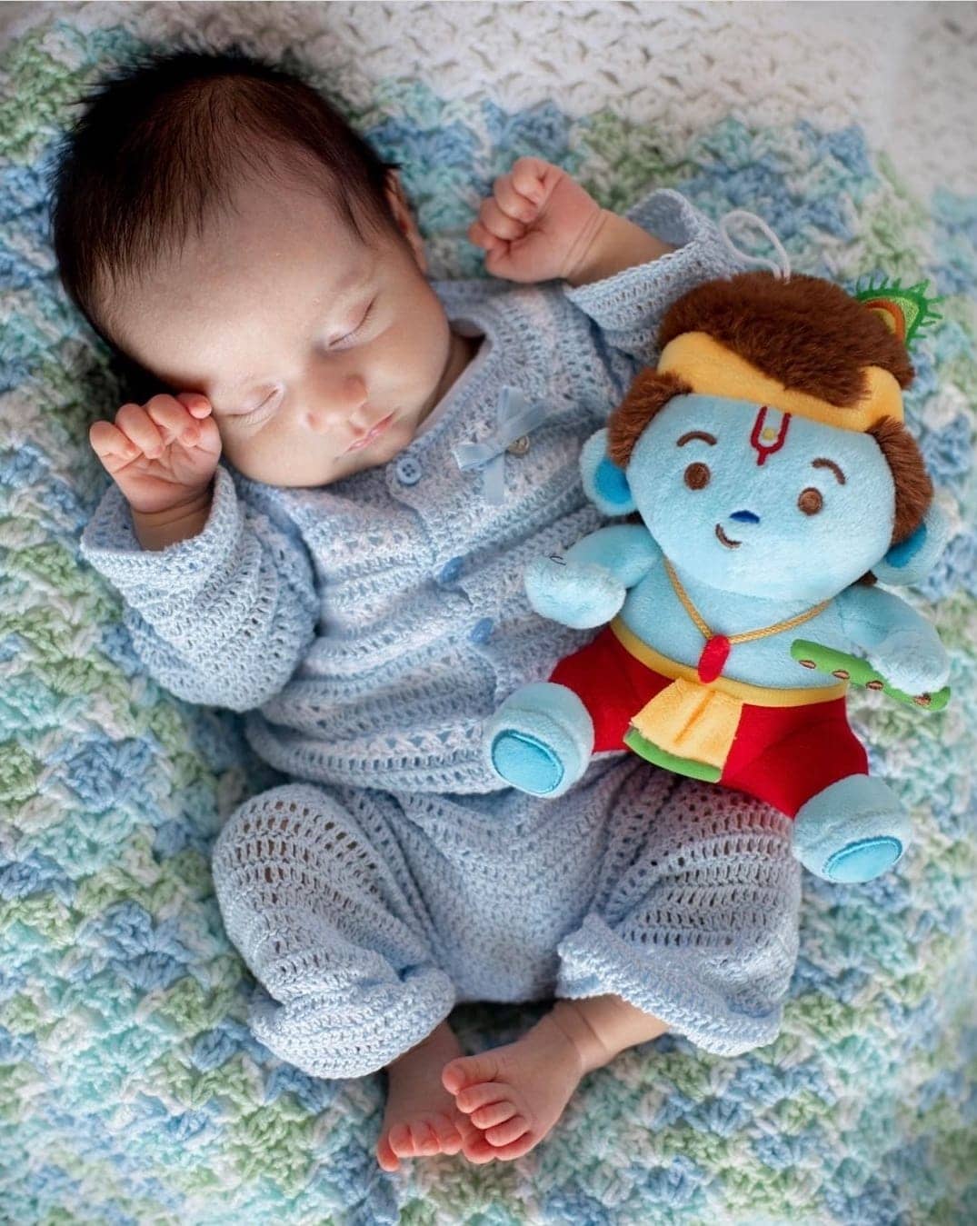 Baby in a blue outfit next to Modi Toys Krishna plush on a patterned blanket.